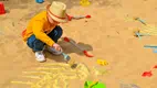 Young toddle wearing a yellow hate and a mustard color sweater with jeans holding a blue brush, uncovering fake fossils in a sandbox