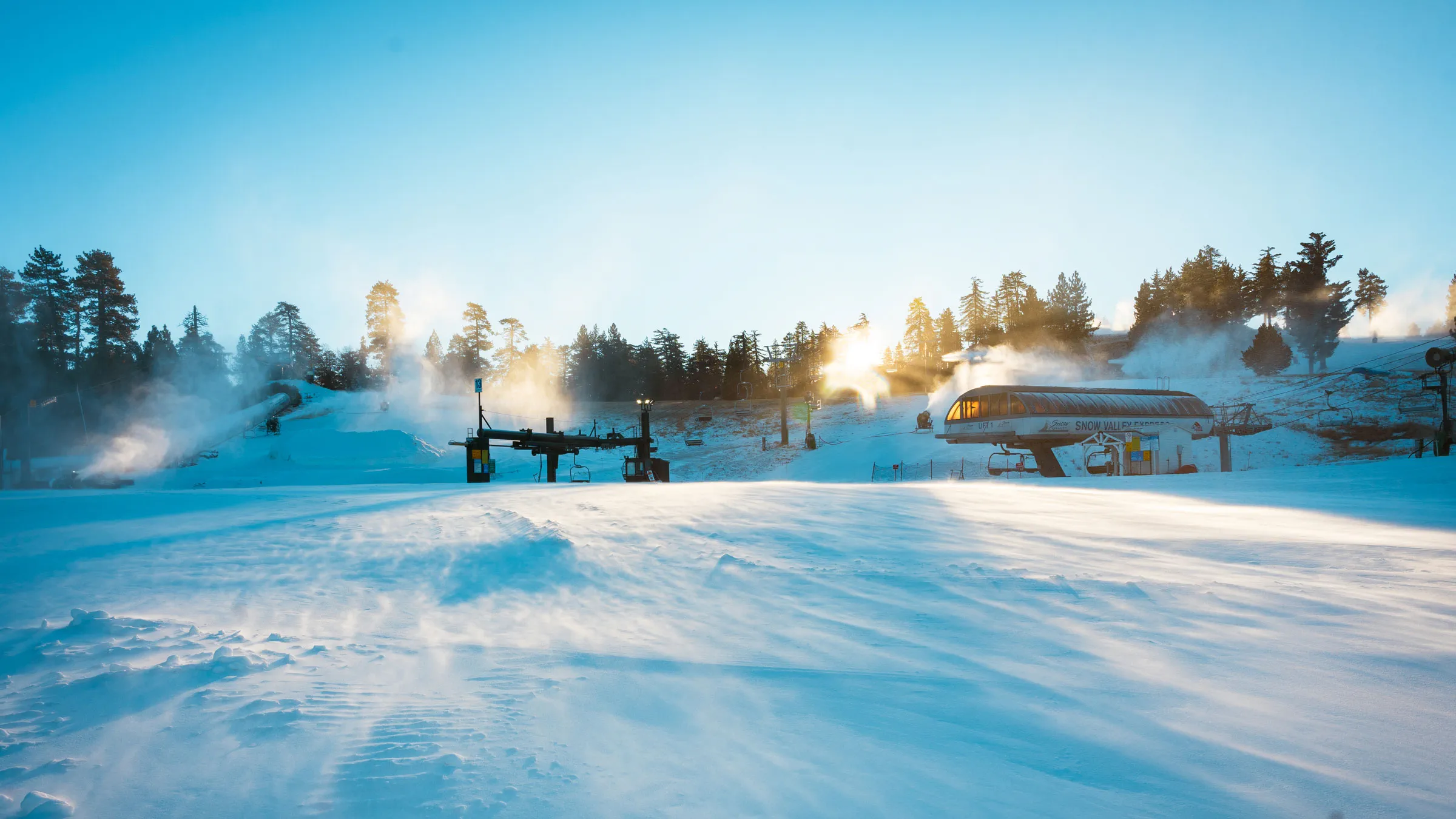 Early morning at Snow Valley, in the base area near chairlifts, with the sun creeping up over the forest trees and snowmaking guns turned on, blowing snow on the mountain trails.