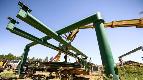 A forklift at the base of Bear Mountain lifting and removing a green chairlift part during a lift overhaul project during the summer, as dirt and green grass lays in the foreground.