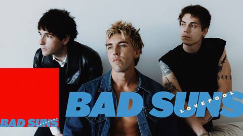 Three adults looking to their right posing for the camera with their band name, Bad Suns, in blue font overlaying the image.