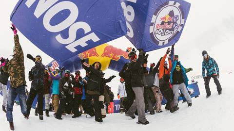 A crowd of people dressed in snow clothing on the snow at a ski resort holding two big blue and white Red Bull dice during an event.