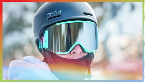 A close up face shot of an adult in a black Smith helmet with turquoise goggles during a winter day