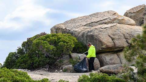 A Big Bear Mountain Resort employee in a yellow jacket with a trash bag picking up trash near a rock formation at Snow Valley during Trash Clean Up Day 2024