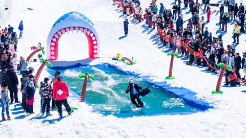 A snowboarder in a suit holding a brief case completing the pond skim at Bear Break 2024