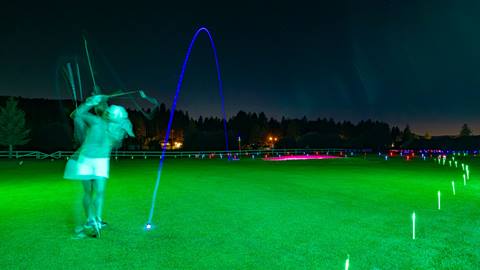 Adult golfer at night chipping a blue glow ball along a glow in the dark lit up fairway onto a putting green with glow lights and a purple glow bunker