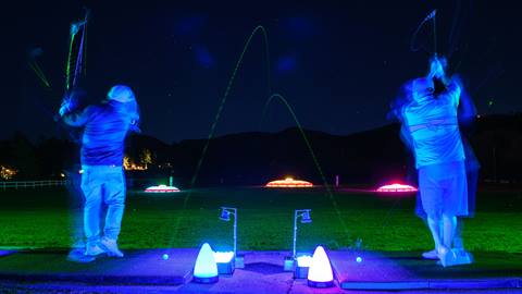 Two golfers, at night, side by side on different practice mats at the Bear Mountain Golf Course Driving Range hitting glow balls to the lit up targets