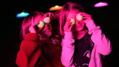 Two kiddos under glow lights at the Bear Mountain Golf Course Driving Range holding glow golf balls up to their eyes and smiling for the camera