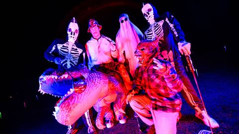 Six golfers at night at Bear Mountain Golf Course dressed up in Halloween costumes ranging from a warewolf, two skeletons, a ghost, and a dinosaur.