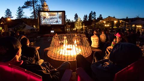 A movie on the big screen in the base area at Snow Summit during a Movies in the Meadow night with crowd goers warming up near a fire pit