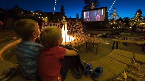 Two kiddos sitting next to a fire pit watching a movie on the blow up big screen during Snow Summit's Movies in the Meadow