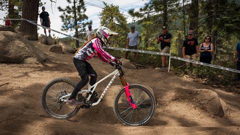 A mountain bike rider in a full face helmet, white and pink jersey, and black pants on a white and pink bike ride on a dirt trail as spectators watch during a Snow Valley race event.