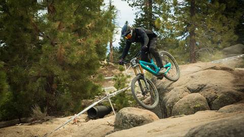 A mountain biker in all black on a teal bike rolling through rocks on a dirt trail at Snow Valley