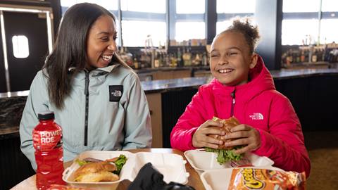 Adult in a light teal jacket and kiddo in pink jacket sitting down inside the main lodge eating lunch that includes Powerade sport drinks, chips, and burgers.