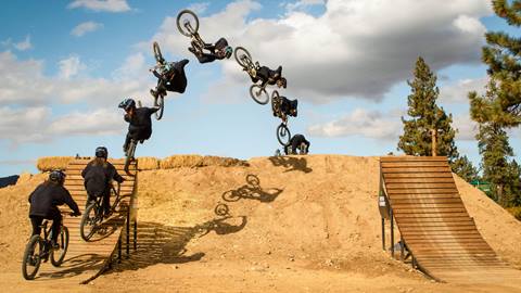 A sequenced shot of professional mountain biker, Dylan Stark, launching off a wooden jump at Snow Summit doing an aerial trick on his bike while landing on a dirt landing.