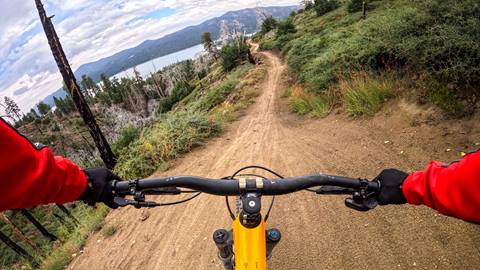 A chest mounted GoPro view of a mountain biker with red long sleeves and black gloves riding a black an orange mountain bike riding a dirt trail with trees and Big Bear Lake views at Snow Summit.