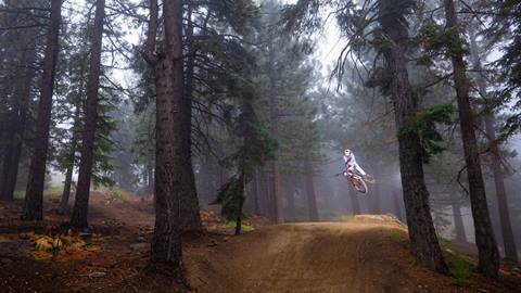 A mountain biker in all white apparel and a full face helmet catching air, while doing a one hander, with trees surrounding the trail at Snow Summit's bike park.