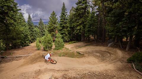 A mountain biker slashing his tire through a dirt berm with surrounding trees at Snow Summit wearing red gloves and white top to bottom apparel.