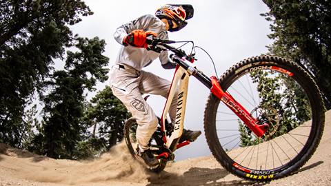 A close up of a mountain biker in a white jersey, white pants, and a black full face helmet on a red and white bike riding on a dirt trail at Snow Summit.