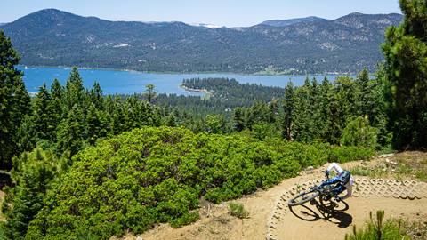 A mountain biker in blue pants, white jersey, and black full face helmet on a paved berm on a Snow Summit bike trail with trees and Big Bear Lake in the background