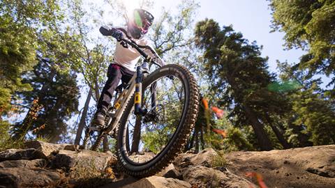 A close up shot of a mountain biker in black pants, white jersey, and black full face helmet with red goggles on their bike rolling through a rock garden on a dirt trail at Snow Summit.