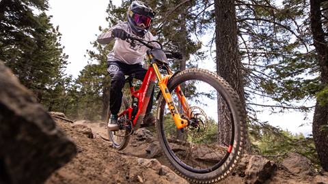 A mountain biker in a black full face helmet, goggles, a white jersey, and black pants on a red bike at Summit Bike Park rolling through rocks with trees lining the dirt trail.