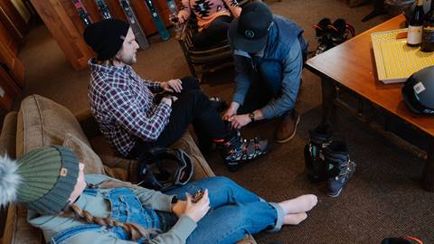 Two adults sitting on a brown coach at their lodging accommodation being sized and fitted with rental ski boots.