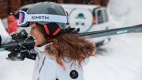 An adult in a white ski jacket, grey helmet, and white Smith goggles holding a pair of rental skis from Ski Butlers over her shoulder while walking on white snow with a white van in the background.