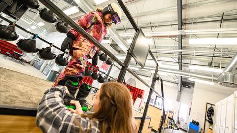 A little kid in a colorful snow outfit and helmet getting fitted in the Rental Shop with snowboard boots