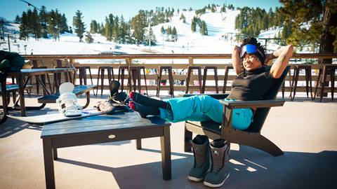 Adult lounging on the Snow Valley sun deck during winter with snowboard on a table, boots on the floor, and feet up relaxing during a bluebird day